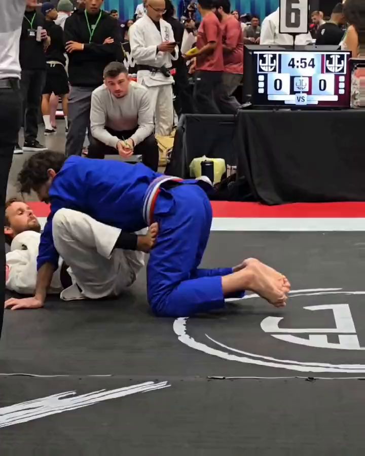 A blue gi passing a white gi's guard at a jiu jitsu tournament, the scoreboard behind