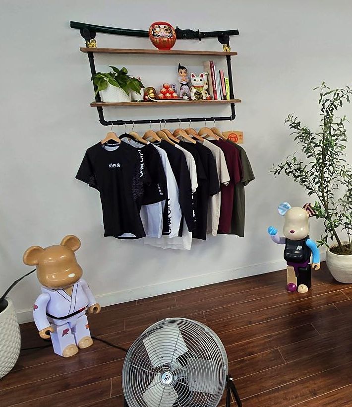 KŌBŌ tees and rashguards on the lounge merch rack under the daruma shelf