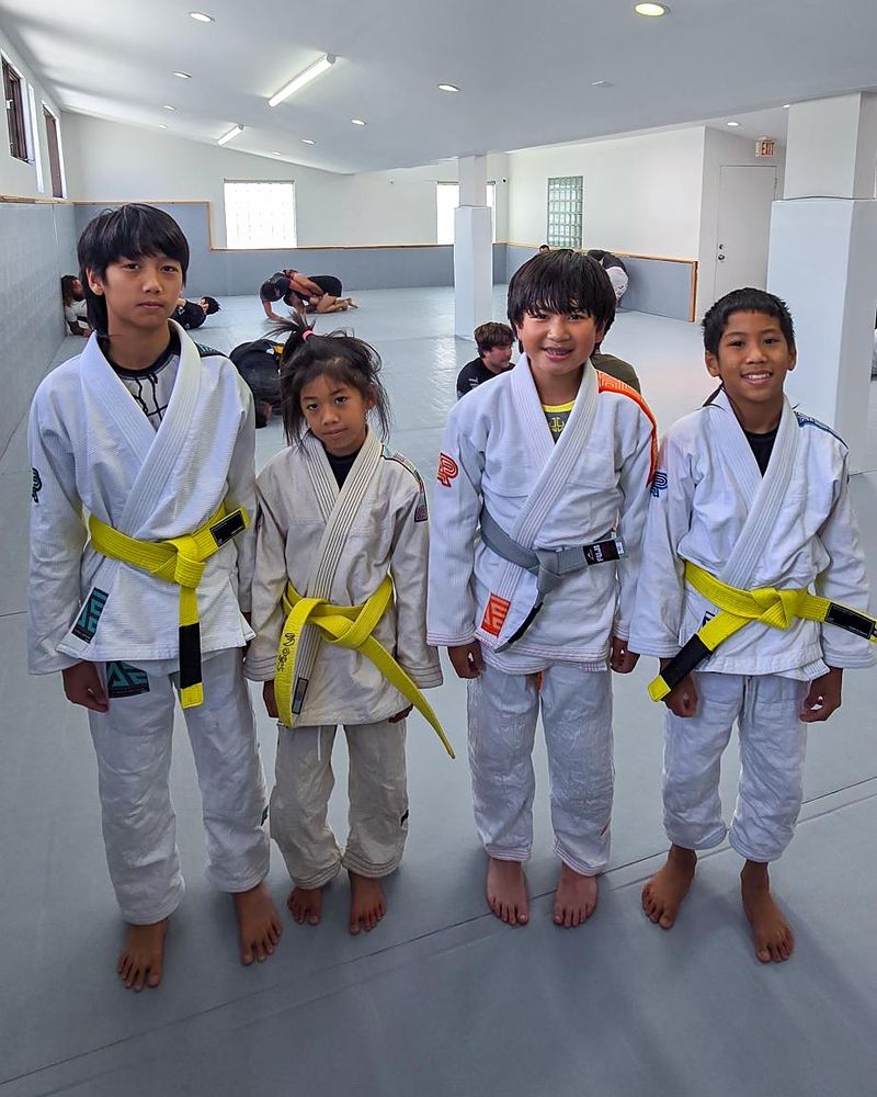 Four kids in white gis with yellow and grey belts, smiling on the mat
